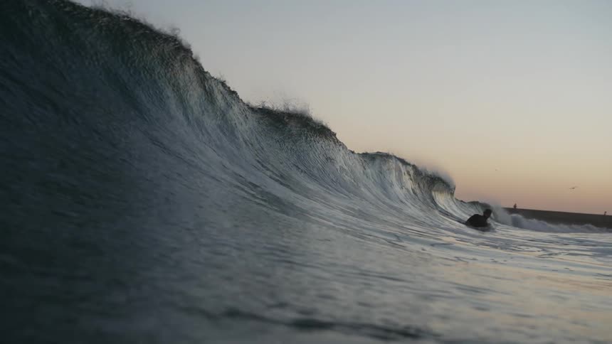 An ocean wave moves toward swimmers in slow motion at sunset