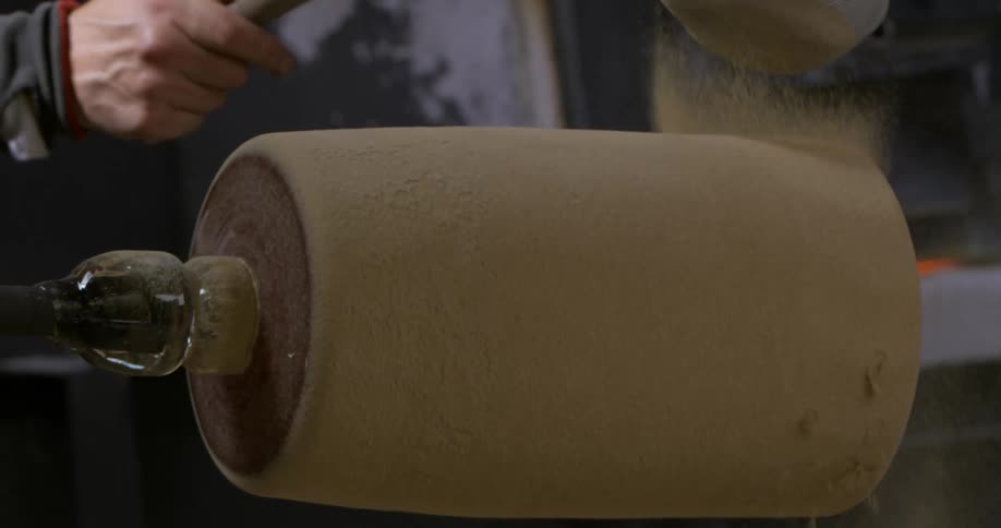 Person coats rotating glass cylinder with sand in workshop