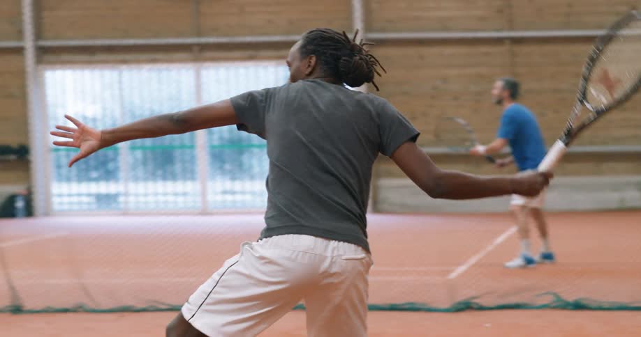 A man swings a tennis ball with a racket on an indoor tennis court while another man holds a racket on another court in the background