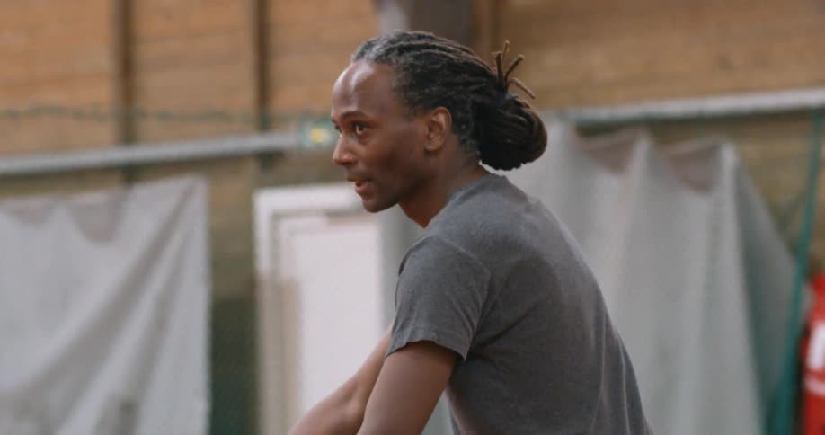 A black man with dreadlocks throws a ball into the air and hits it with a tennis racket
