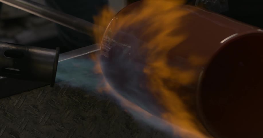 An opaque jar rotates on a machine while a tool blows fire at it