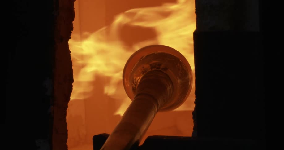 A long rod with a circular object attached to it rotates in a kiln with fire blowing on it