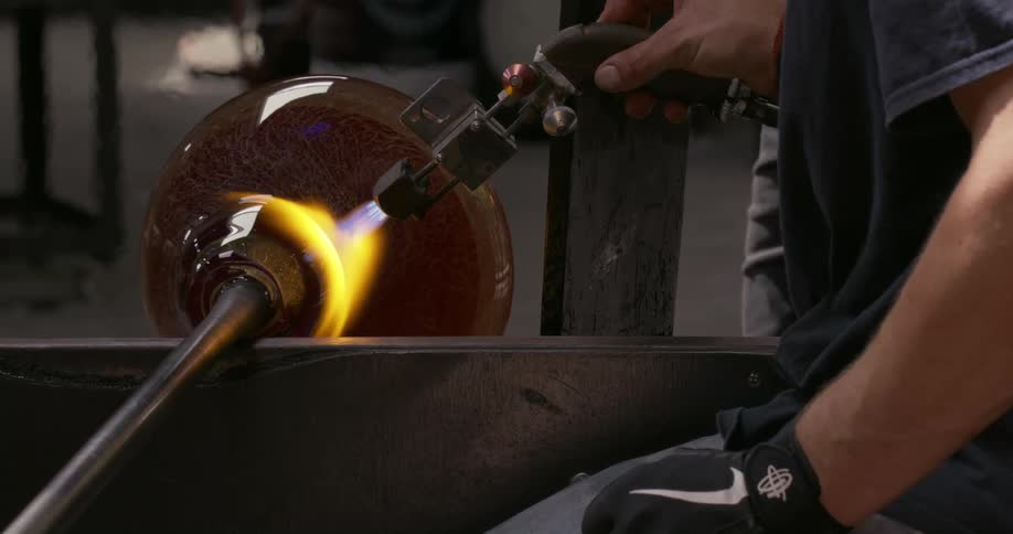 A man holds a torch and blows it on a jar that rotates on a metal rod