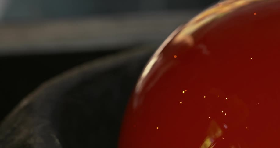 Smoke rises from metal container as molten glass sphere rotates on stick during glass making