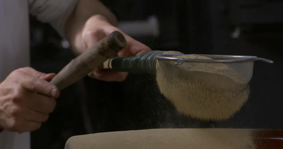 Glass maker taps sieve with stick to coat rotating glass cylinder with sand