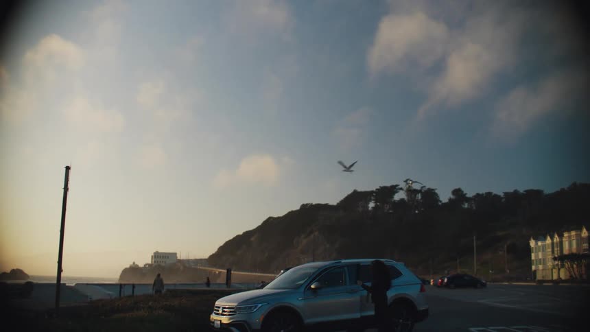 Camera pans down from blue sky and clouds to show a woman getting out of a car that is parked along the coast closing the door as she walks away toward the shore birds fly overhead