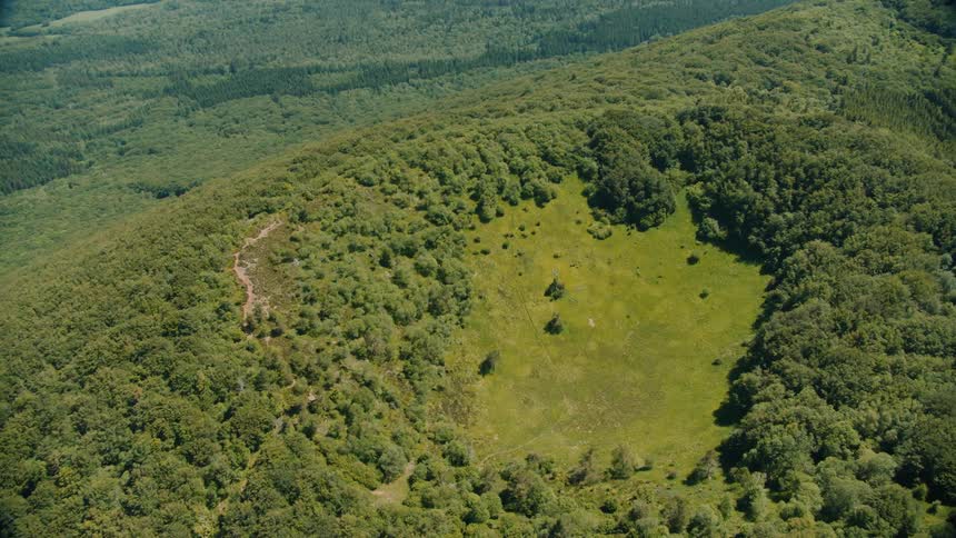 A aerial shot of a grassy area on top of a mountain the grassy area is circular surrounded by green trees