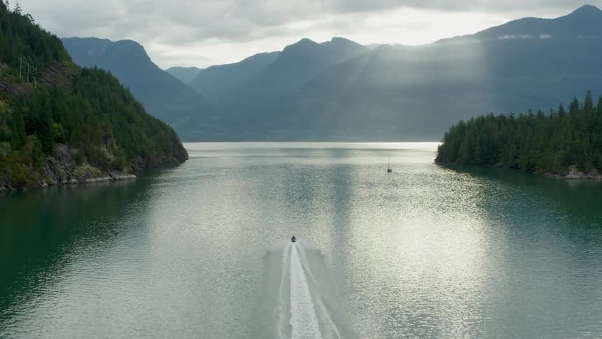 Jet ski leaves wake as it nears sailboat between forest covered shores on foggy day