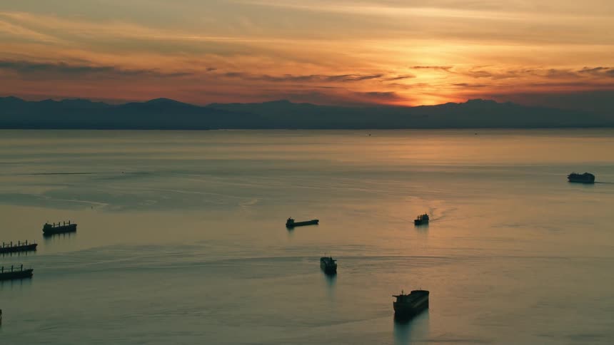 Multiple boats sit on water as setting sun silhouettes mountains on shore