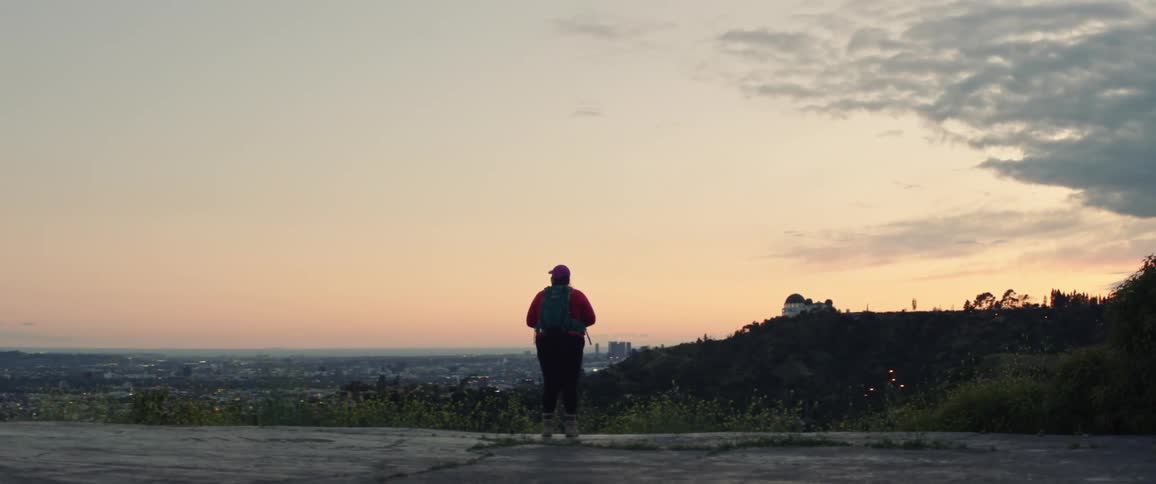 Woman with a cap a backpack leggings and hiking poles watches city skyline at sunset