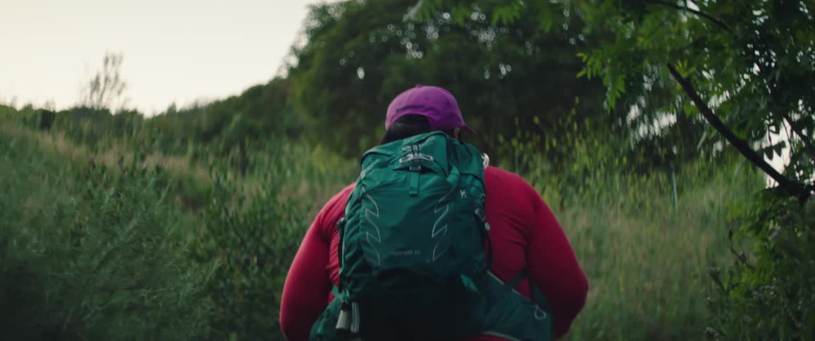 Woman wearing a purple cap a red jacket and a green backpack walks uphill