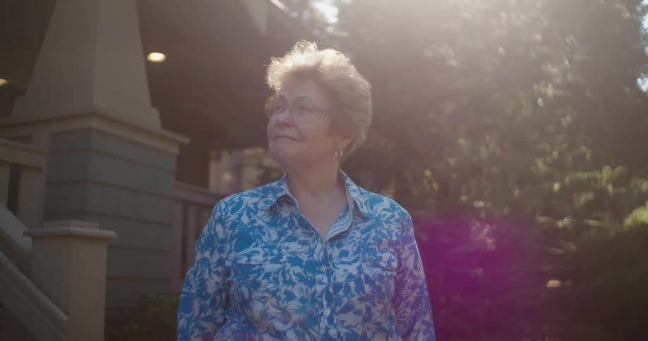 An elderly woman wearing glasses stands in front of a house and looks up as her hair is illuminated by sunlight