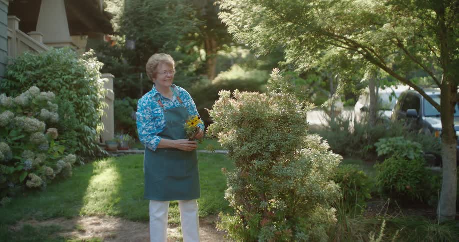 Older woman clipping flowers in her front yard waves and smiles