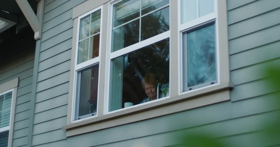 Exterior of a house and an elderly woman wearing blue shirt looking out the window