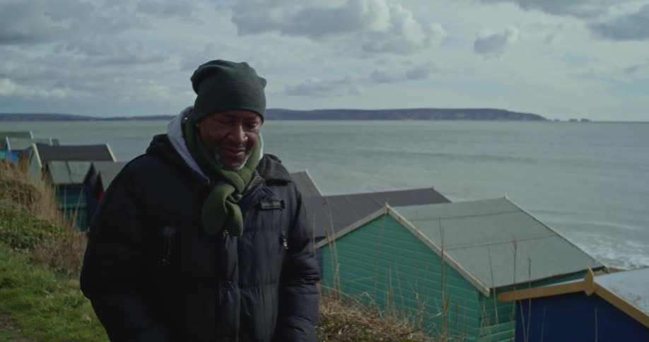 Man in a  coat walks on a path, through colourful huts, near the sea