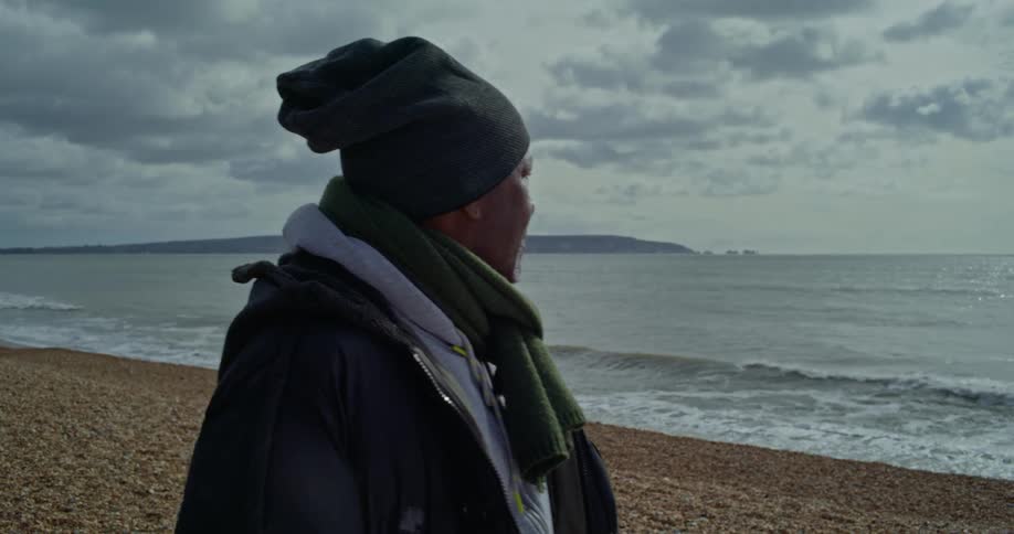 Older man wearing cold weather gear walks along beach and looks out to sea on a cloudy day