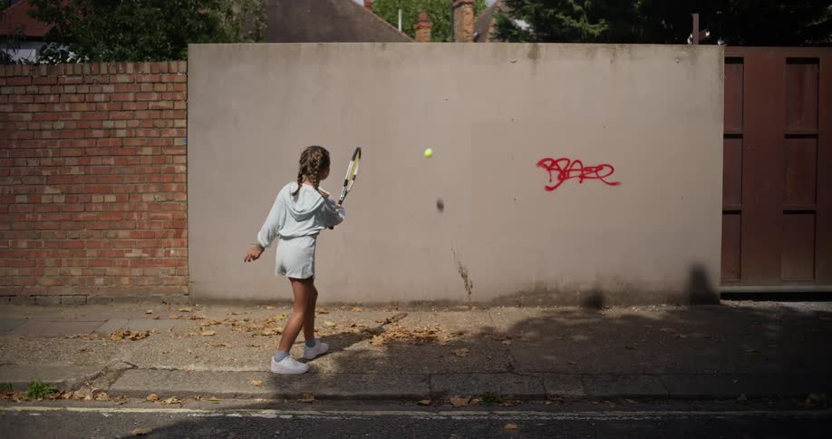 Girl with braids a light blue hoodie and shorts plays tennis against the wall