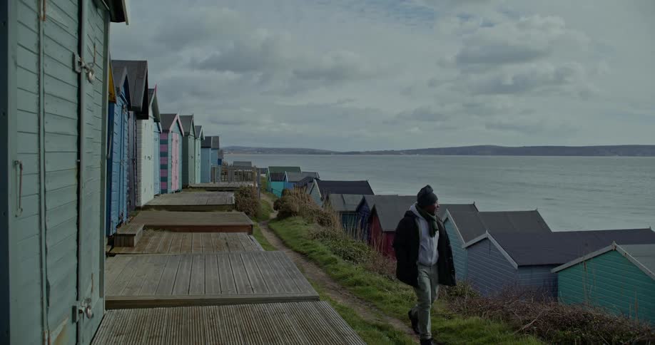Man walks on a path through colorful wooden beach huts overlooking the sea