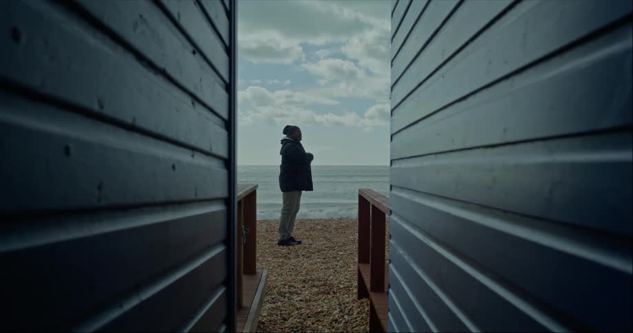 Man with a black hat and coat walks along pebble beach