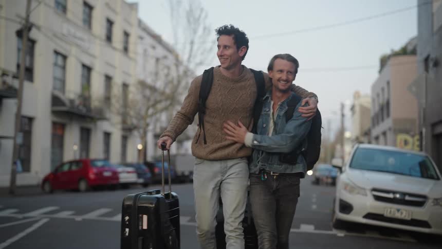 Two men are walking on a city street pulling their luggage and talking as they walk past cars and buildings