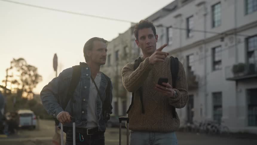 Two men are standing on a downtown street looking at a cell phone as one man points and they pull their luggage and walk away