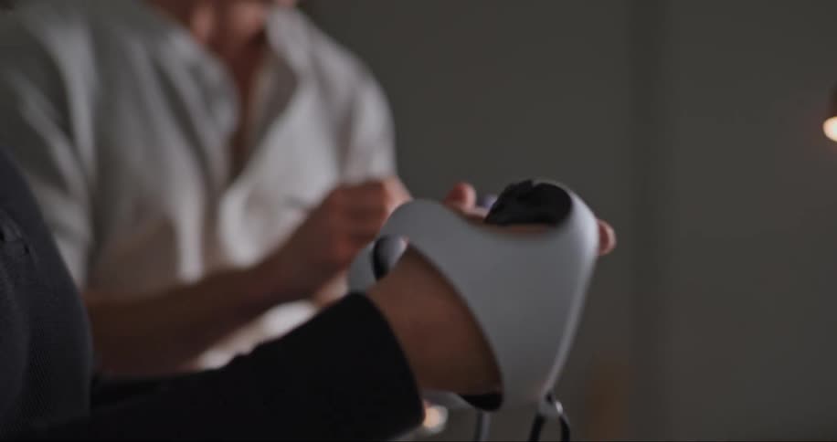 A man is sitting down as he wears a virtual reality headset looking around moving controllers in his hands as a man standing next to him writes on a tablet