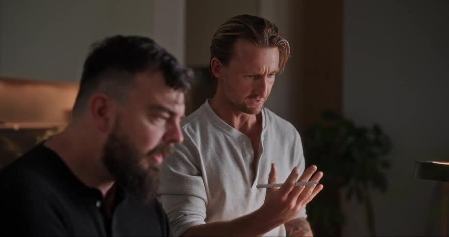 A man is standing with a tablet and a pen in his hand and points as he talks to a man sitting next to him