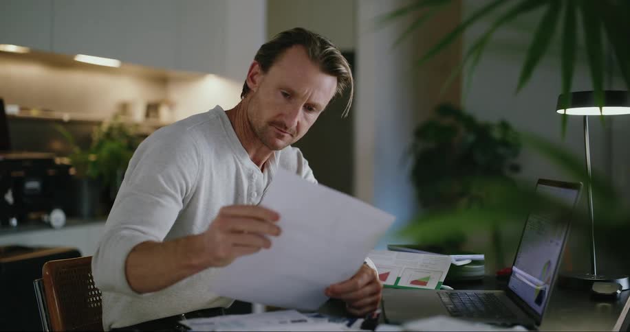 Man sitting at a desk reads a paper while working on a laptop