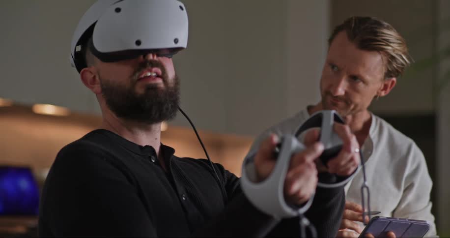 A man is sitting down as he wears a virtual reality headset looking around with controllers in his hands as a man standing next to him holding a tablet