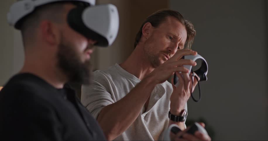 A man is wearing a virtual reality headset as he takes a controller off his wrist and a man standing next to him looks at it as they talk