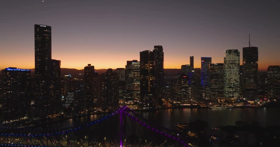 Lights on towers and skyscrapers glow on the coastal city skyline as traffic moves over the bridge and the orange sky colors the horizon