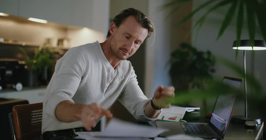 A man is typing on his laptop and looking at papers