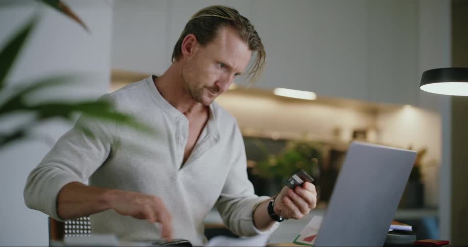 A man is sitting at table with a laptop computer looking at a document and he takes a credit card out of his wallet and types on the keyboard