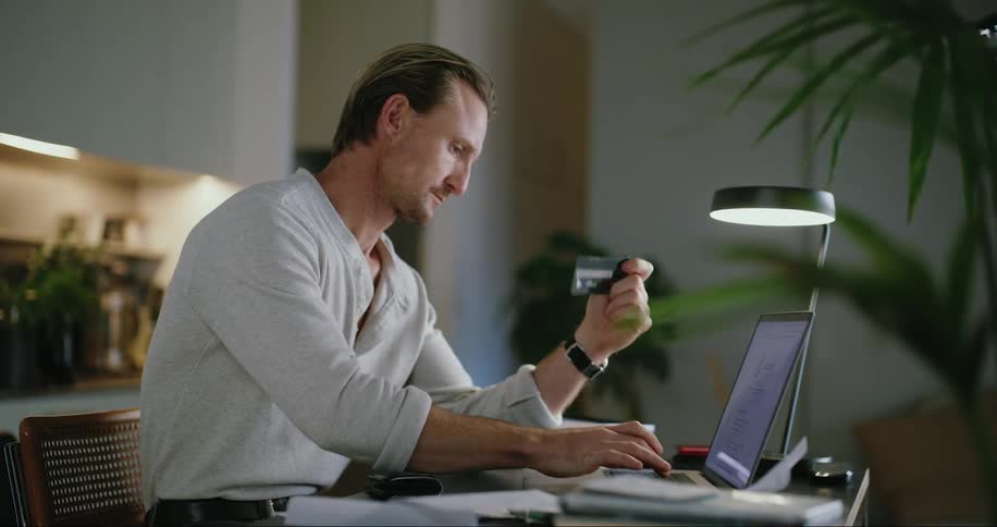 A man sitting in front of his laptop takes his card from his wallet types it into the computer and then crumples a piece of paper and tosses it