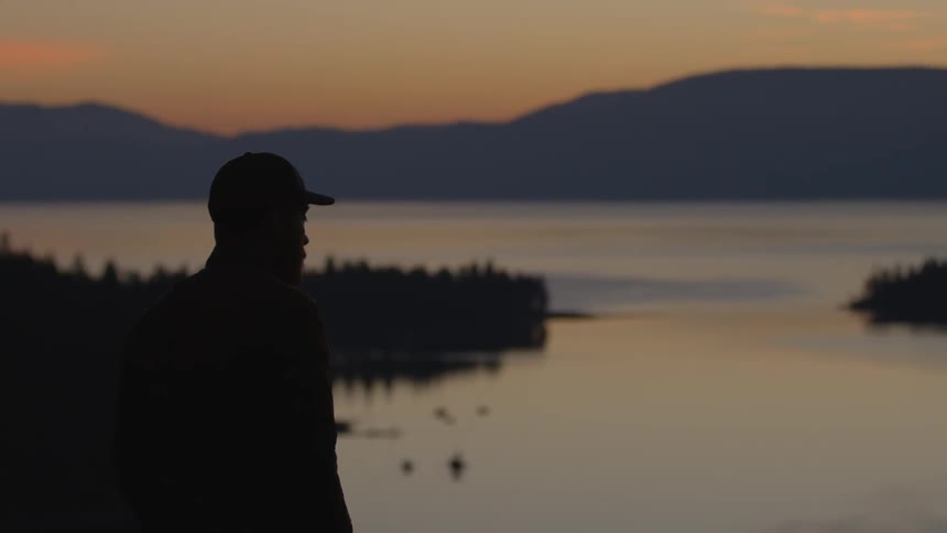A man stands before a lake watching the sunset