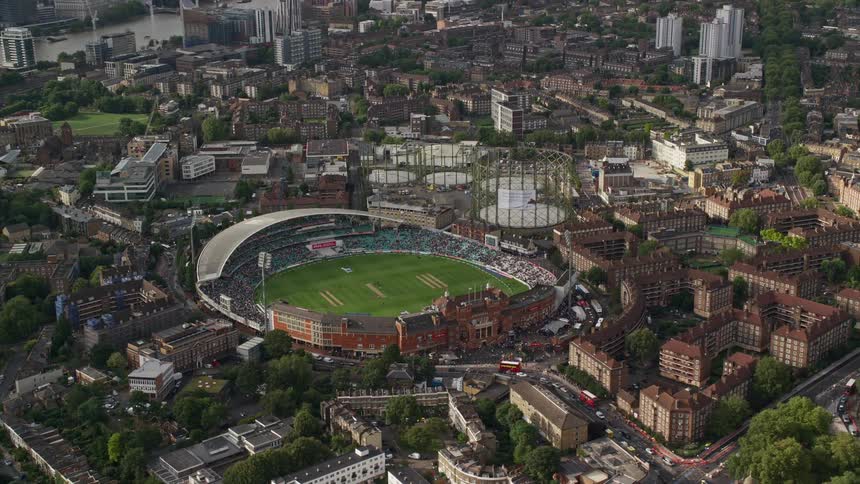 A stadium with a green field nestled within a city nearby there are numerous residential buildings lining a road and vehicles can be seen passing by