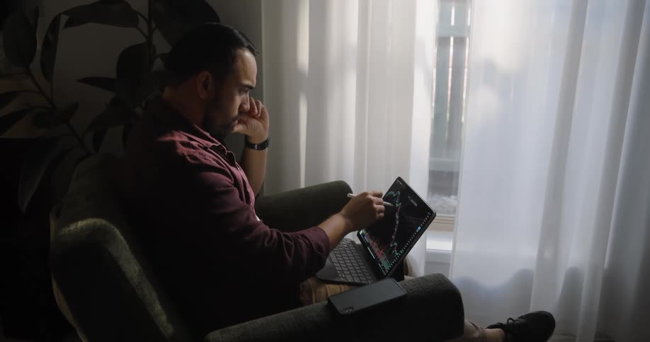 A man on a couch is using a laptop to work on a stocks chart