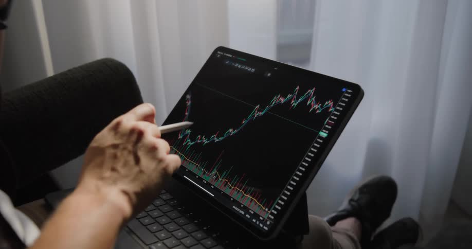 A man points and draws on a graph on a laptop while sitting in a room next to a window