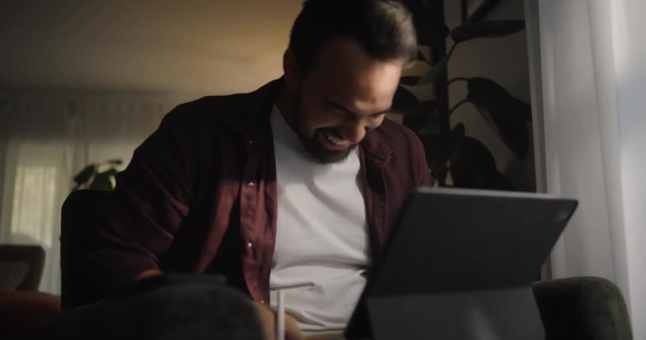 Man with beard white and red shirt sits in armchair using tablet and laughing