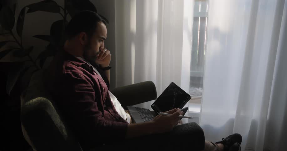 Man sits on a chair next to a window working on a laptop