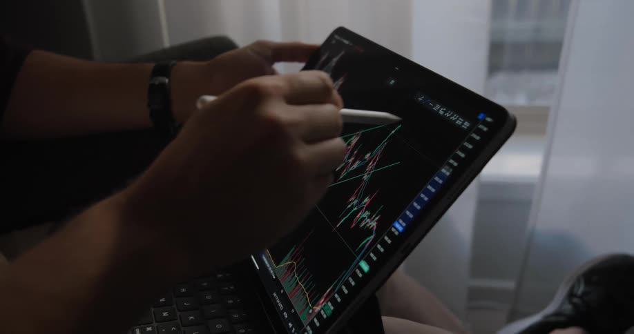 Hands of a person holding a digital pen and sketching on the screen of their tablet with day trading charts