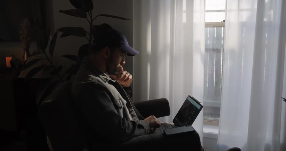 A man is sitting in a living room chair near a window typing on a laptop computer