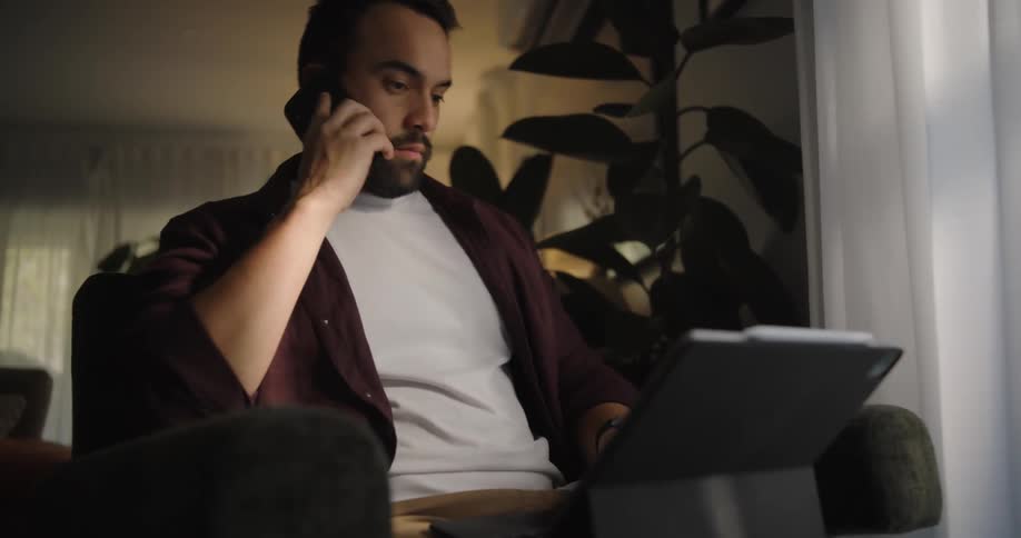 A man is sitting in a living room chair typing on a laptop computer as he answers his phone