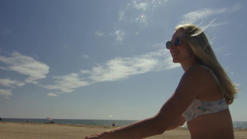 A woman is riding a bicycle wearing sunglasses and a bikini top and jean shorts next to a beach