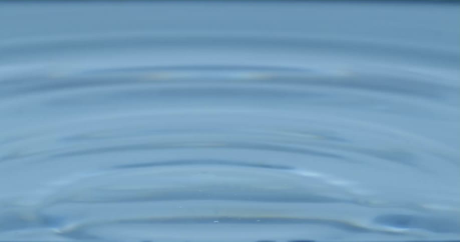 A droplet falls into the water as it splashes upward and down and back up again as ripples spread across the surface