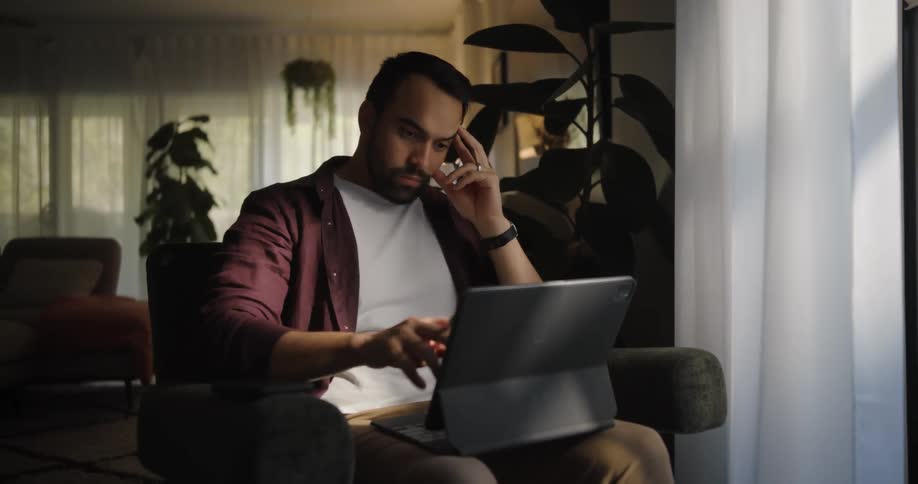 A man sitting on his couch in a darkened room works on a laptop until he gets frustrated and flips his pen