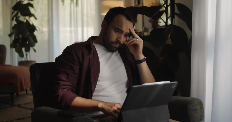 A man on a couch with a laptop on his lap is frustrated with his work