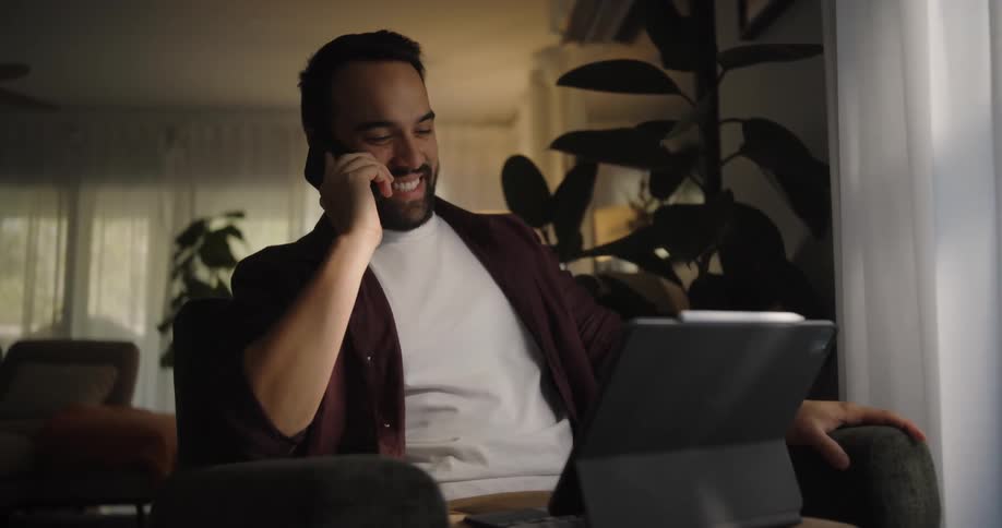 A man is sitting in a living room chair typing on a laptop computer as he answers his phone and smiles as he talks