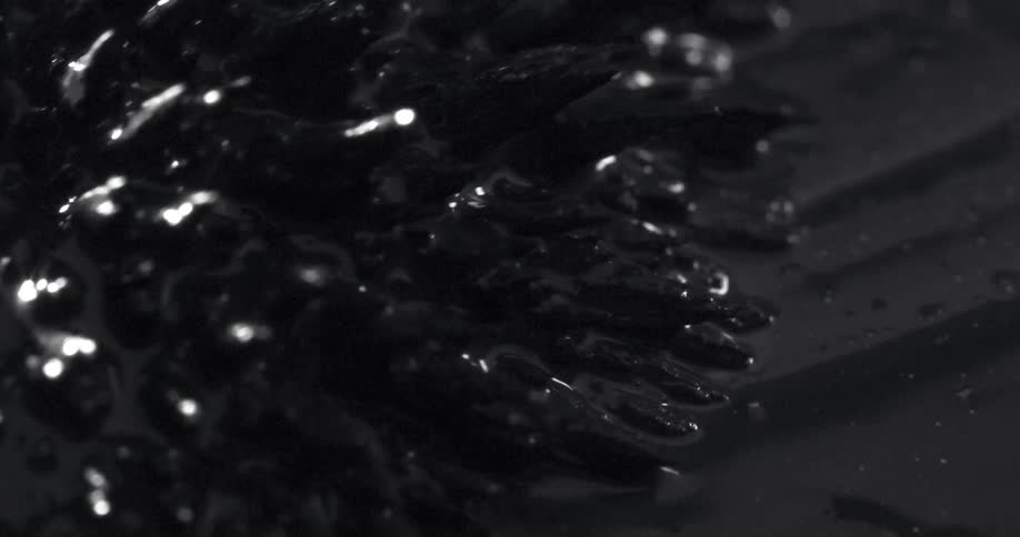 Black object with surface spikes moves across oily black surface before flexing its spikes