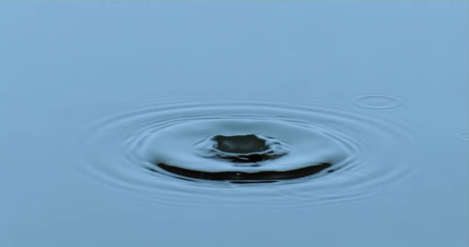 A droplet falls into the water as it splashes upward and down and back up again as ripples spread across the surface